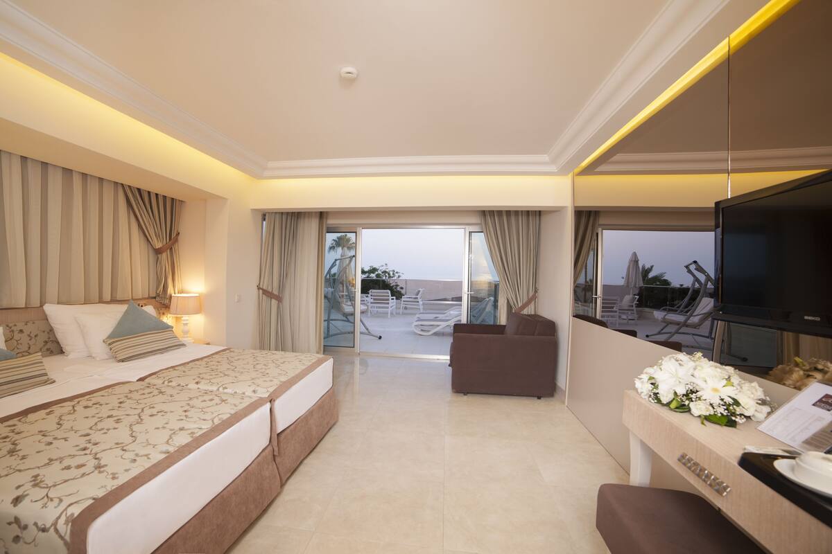 A spacious hotel room with a large bed, a seating area with a TV, and a balcony with a view.