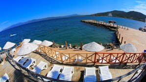Eine Strandterrasse mit Liegestühlen und Sonnenschirmen, ein Steg, der ins Meer führt, und eine gebirgige Kulisse.