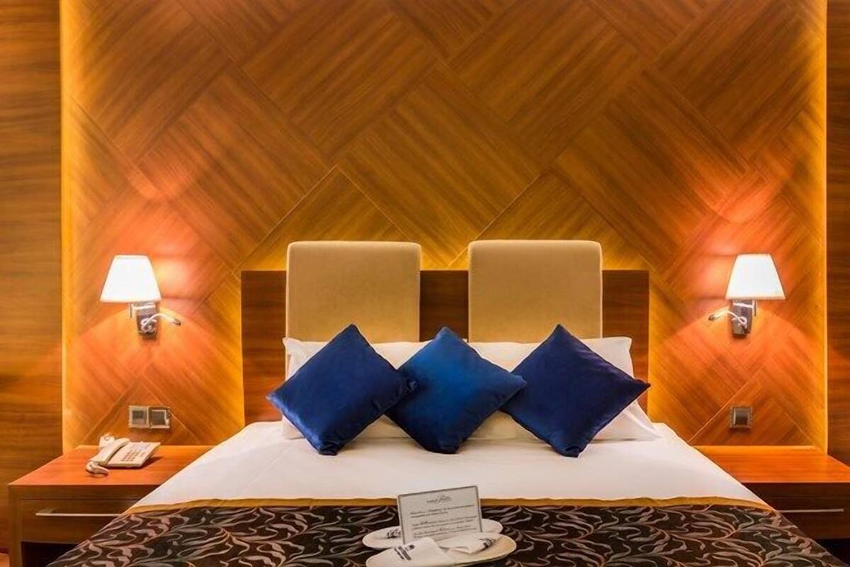 A hotel room with a wooden herringbone wall, a bed with blue pillows, two bedside lamps, and a nightstand with a phone.