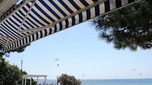 Un espace extérieur aménagé avec des tables et des chaises en osier blanc, offrant une vue sur une plage animée où l’on peut voir des gens et des cerfs-volants.