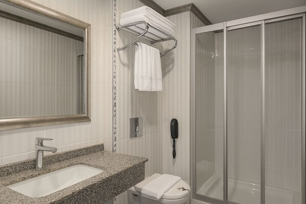 Une salle de bain avec un lavabo surmonté d’un plan de travail en granit, un miroir, un porte-serviettes avec des serviettes, des toilettes et une douche à parois de verre.