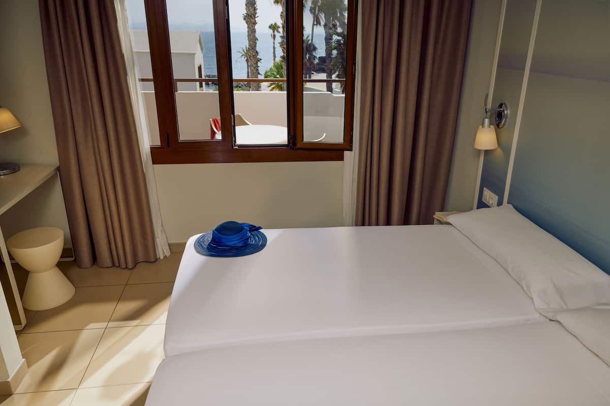 Une chambre d’hôtel avec un lit, une fenêtre donnant sur des palmiers et la mer, une table de chevet avec une lampe, et une serviette bleue sur le lit.