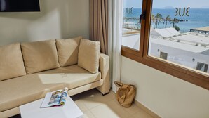 Un sofá color beige, una revista, un bolso tejido y una ventana con vistas al mar y edificios.