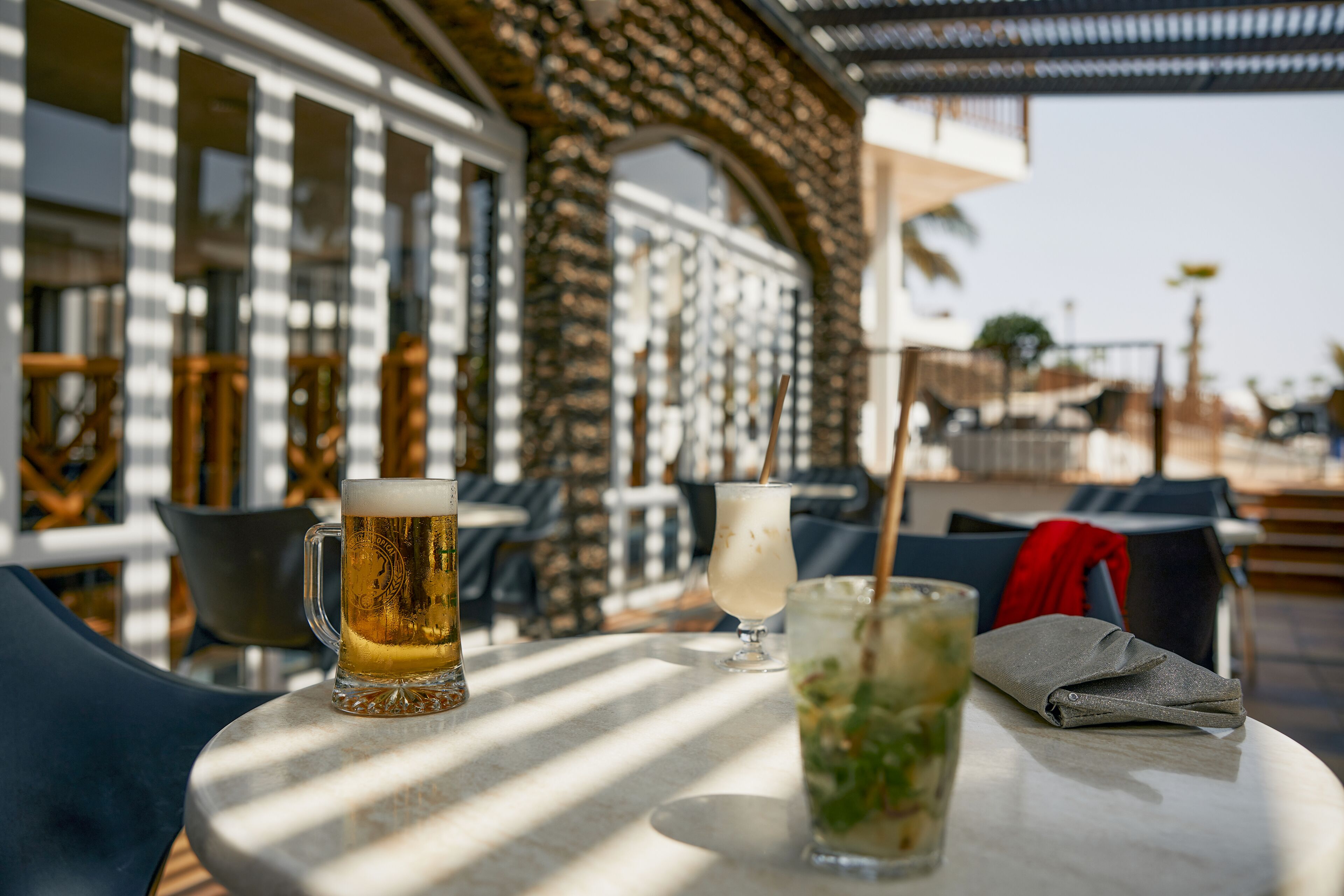 Una mesa con bebidas y una servilleta, ubicada en un área exterior soleada con un muro de ladrillo y grandes ventanales.