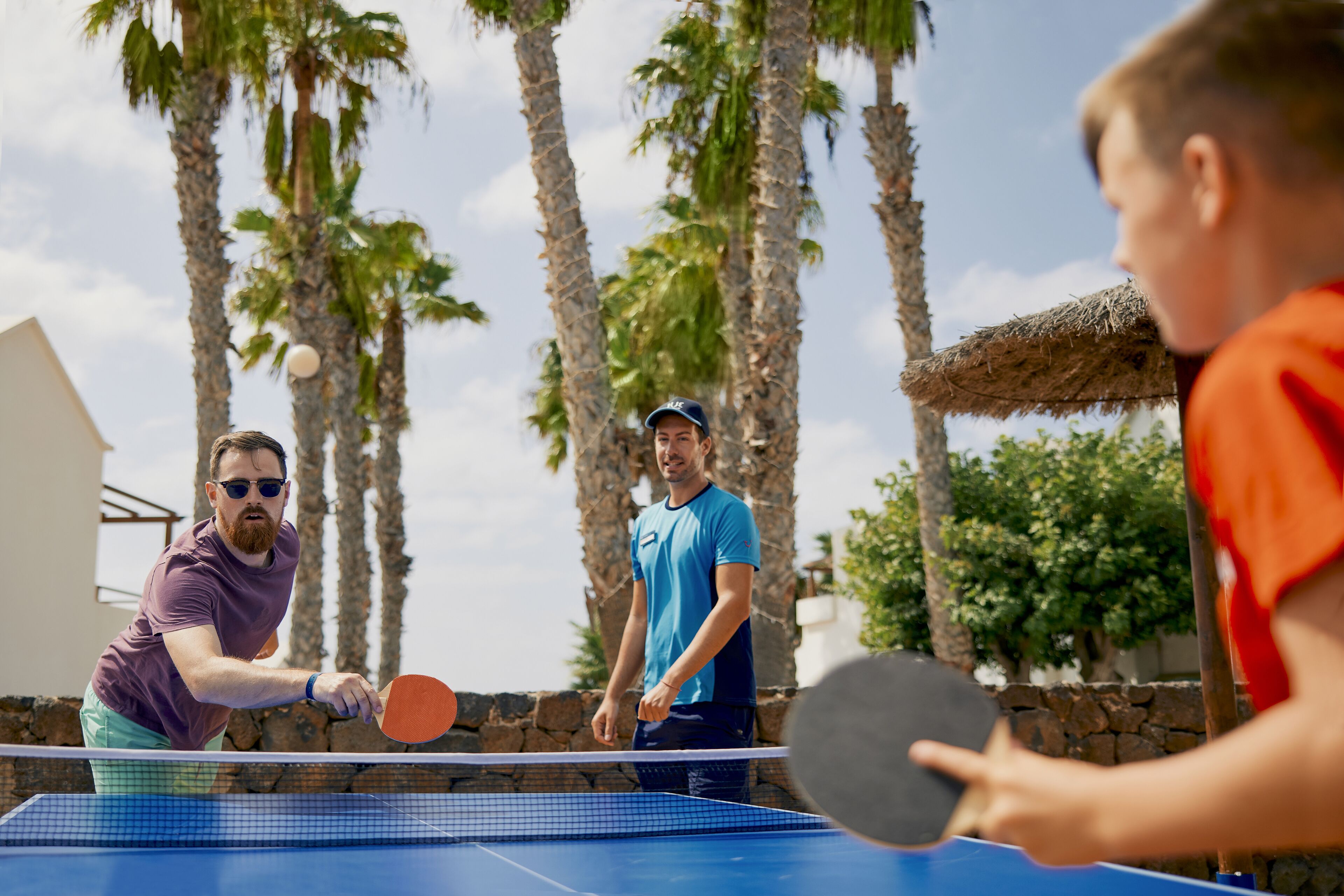 A tennis court with players in action, surrounded by palm trees and a stone wall.