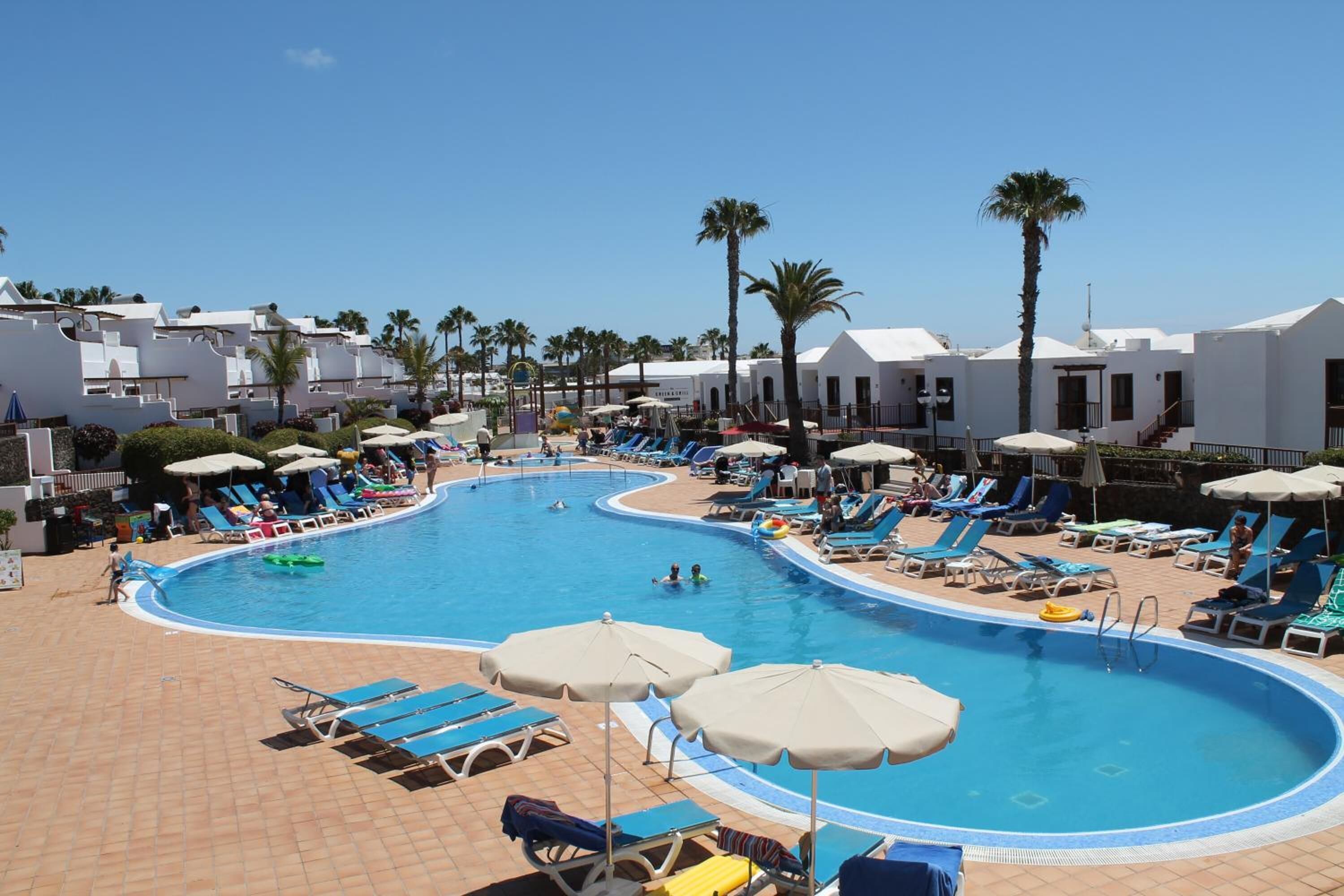 A pool area with sun loungers and umbrellas, surrounded by white buildings and palm trees.