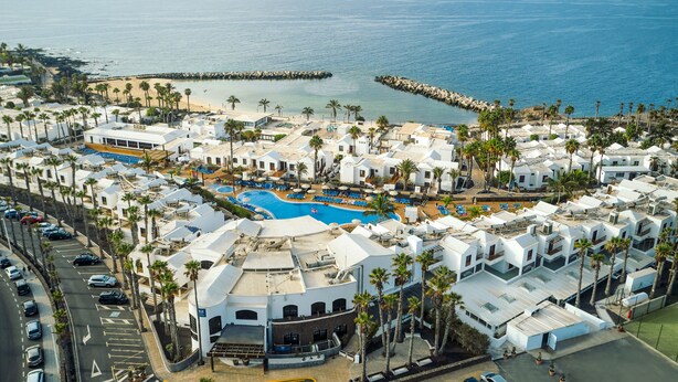 Vista aérea de un complejo turístico frente al mar, con piscina, edificios blancos y palmeras.