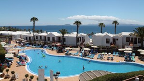 Zona de piscina con sillones de descanso y sombrillas, rodeada de palmeras y vista al mar.