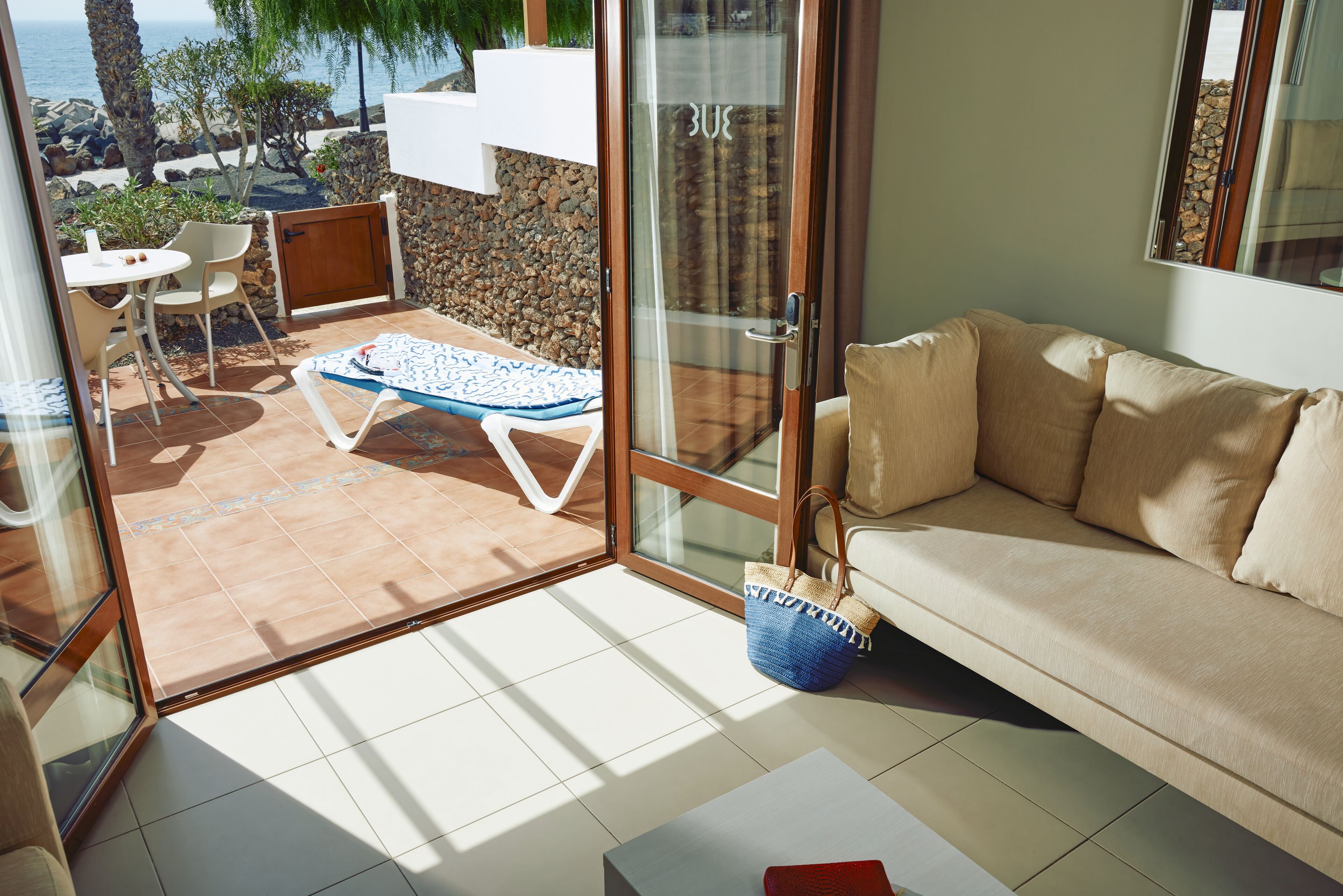A modern living room with a beige sofa, a glass door leading to a patio with a pool, and a view of the sea.