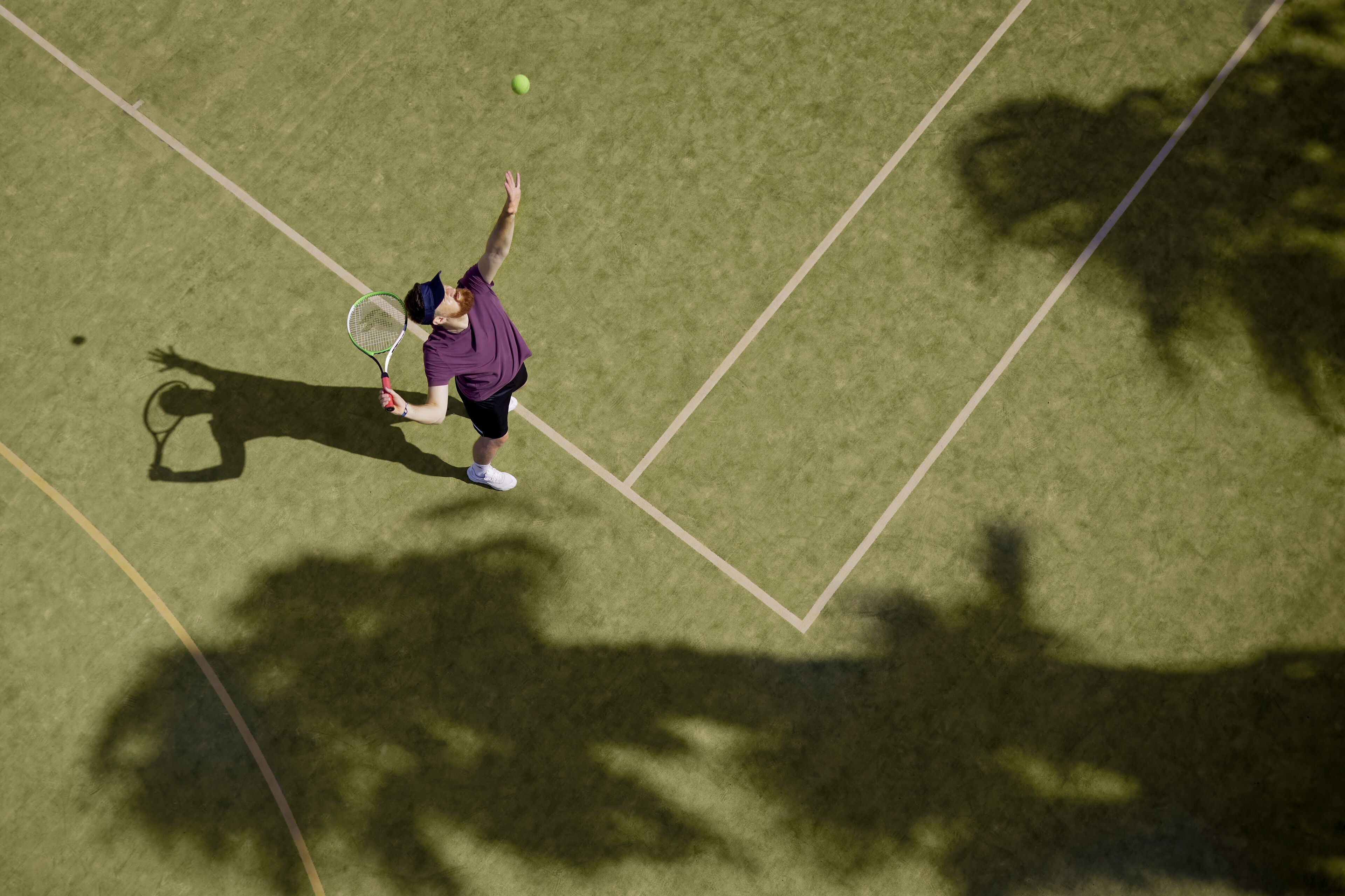 A tennis player in motion on a grass court, with shadows of trees and a camera visible.