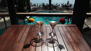 A wooden table with two martini glasses containing drinks, overlooking a pool area with people and tables. - Arora Hotel (Kuşadası)