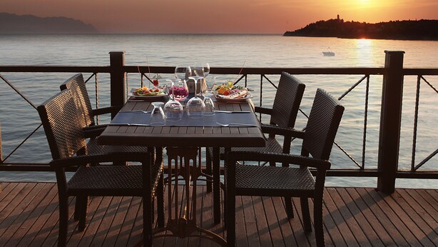 A table set for a meal on a wooden deck with a view of the sunset over the water. - Arora Hotel (Kuşadası)