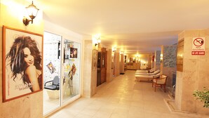 A hotel corridor with a salon on the left, a reception area in the center, and a lounge area with chairs on the right. - Arora Hotel (Kuşadası)