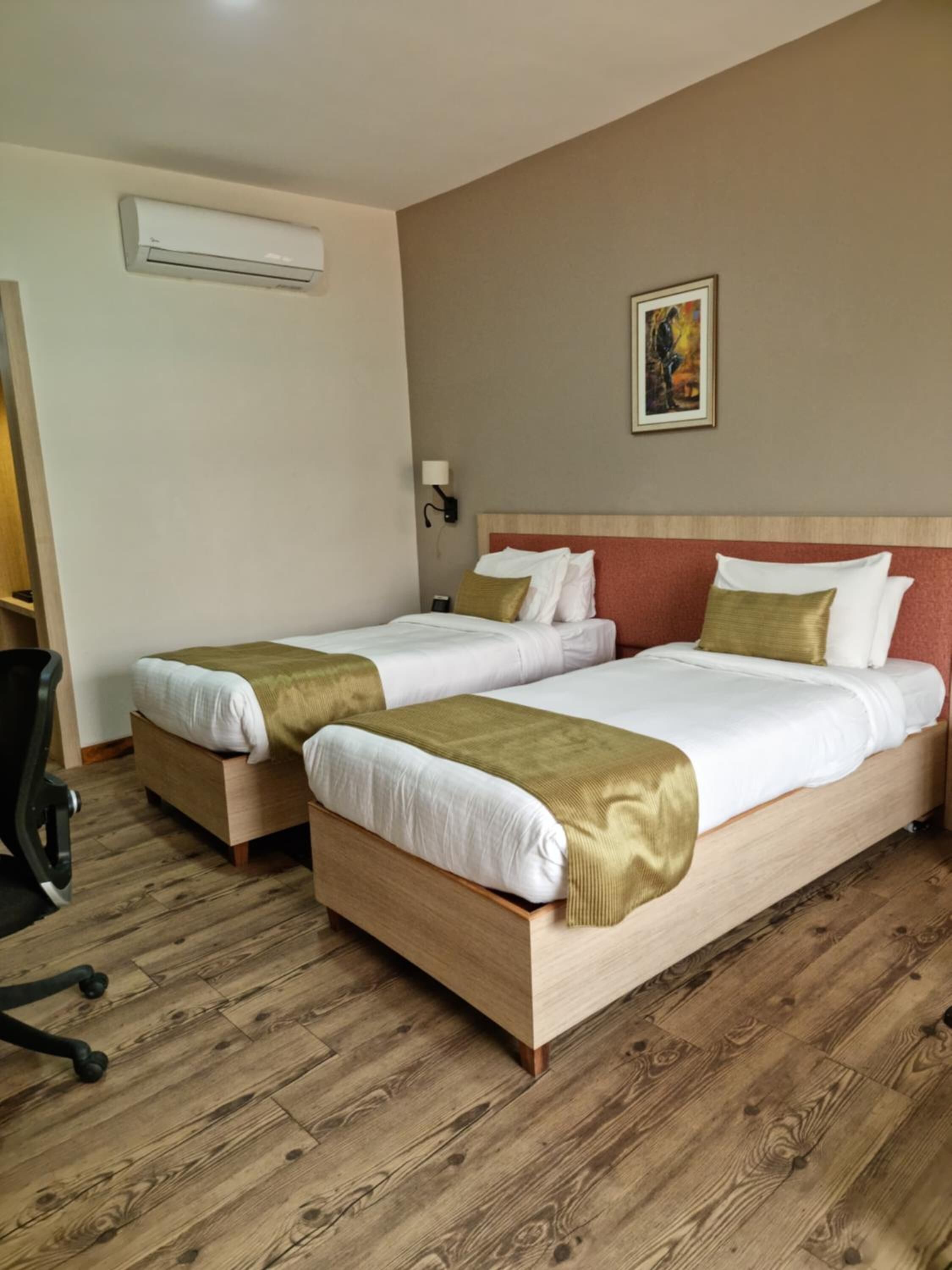 A hotel room with two beds, a wooden floor, a wall-mounted air conditioner, a painting, and a desk with a chair.