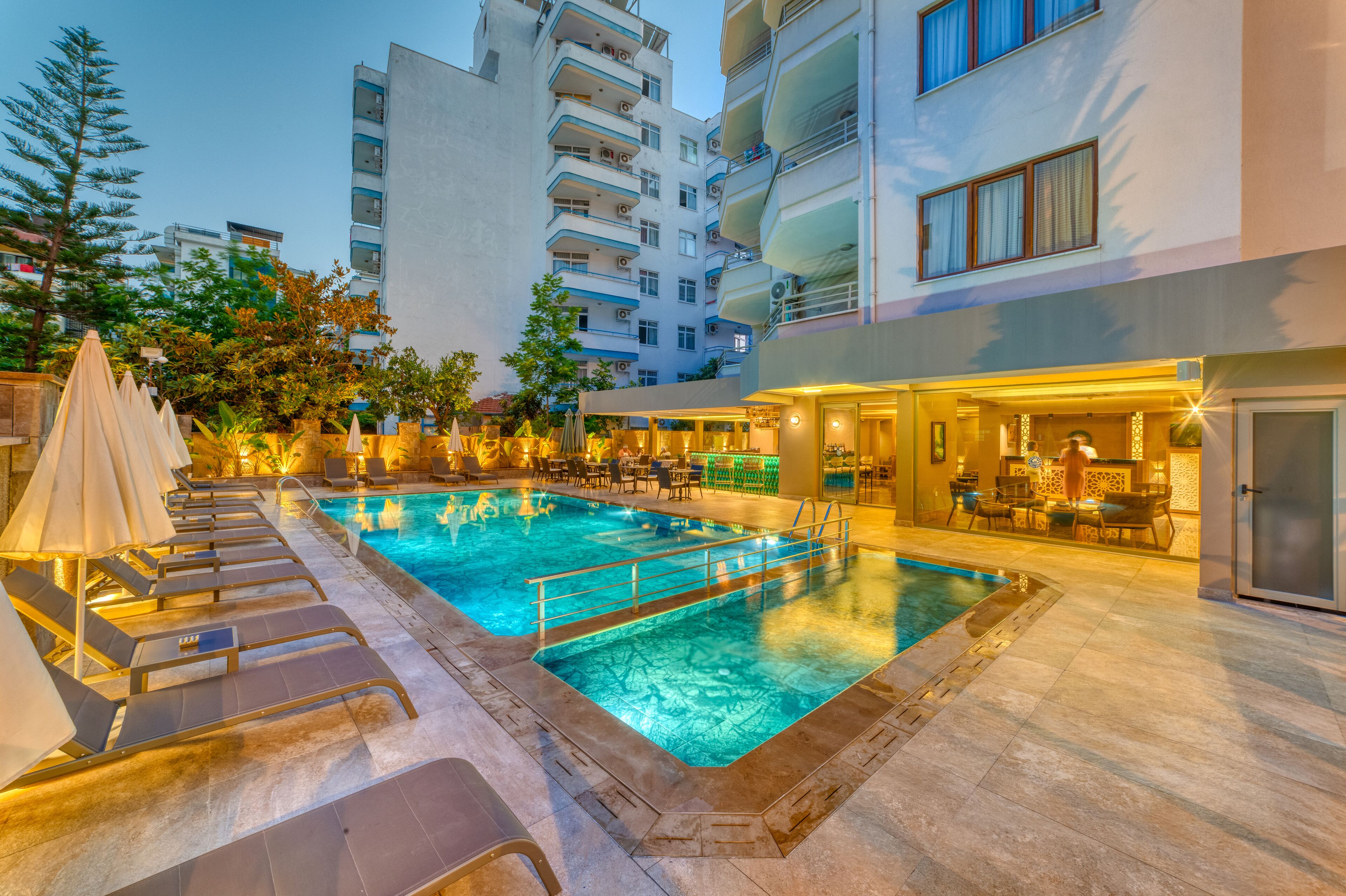 Une piscine avec des chaises longues et des parasols, un espace bar et un bâtiment doté de plusieurs balcons.
