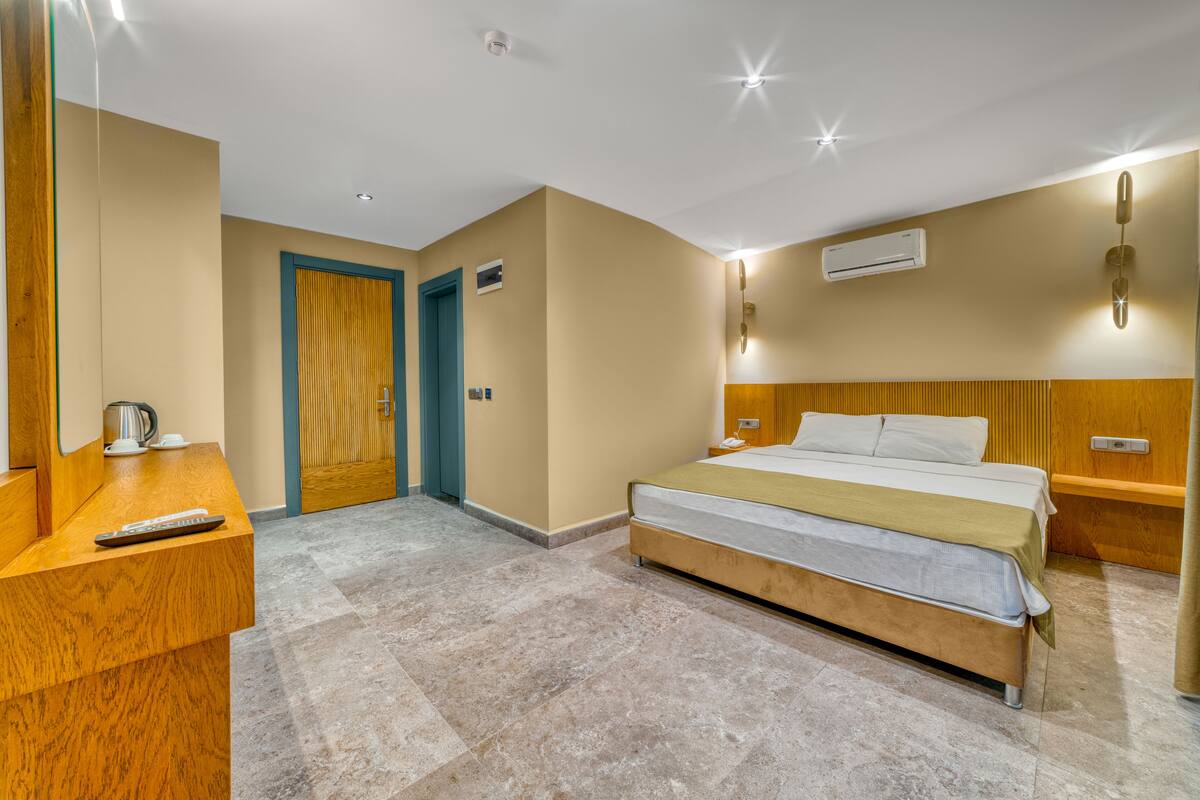 A hotel room with a bed, bedside tables, a wooden desk, a mirror, a door, and an air conditioning unit.