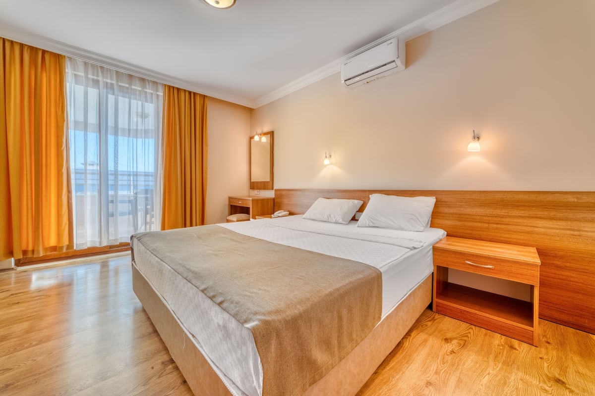 A modern hotel room with a large bed, wooden headboard, bedside tables, and a large window with curtains.