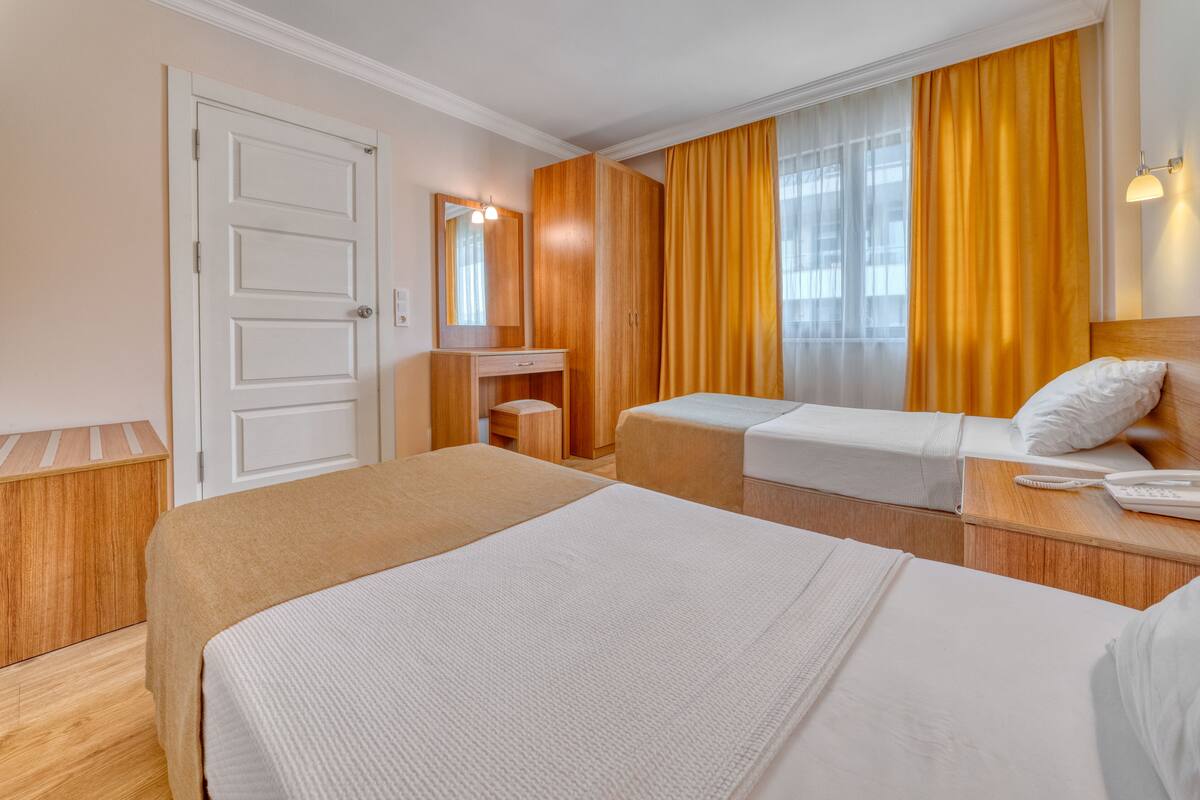 A hotel room with two beds, a wooden wardrobe, a bedside table with a phone, and a window with curtains.