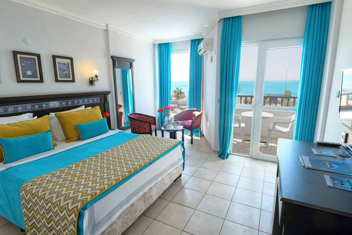 A hotel room with a bed, a desk, a chair, a TV, and a balcony with a view of the sea.