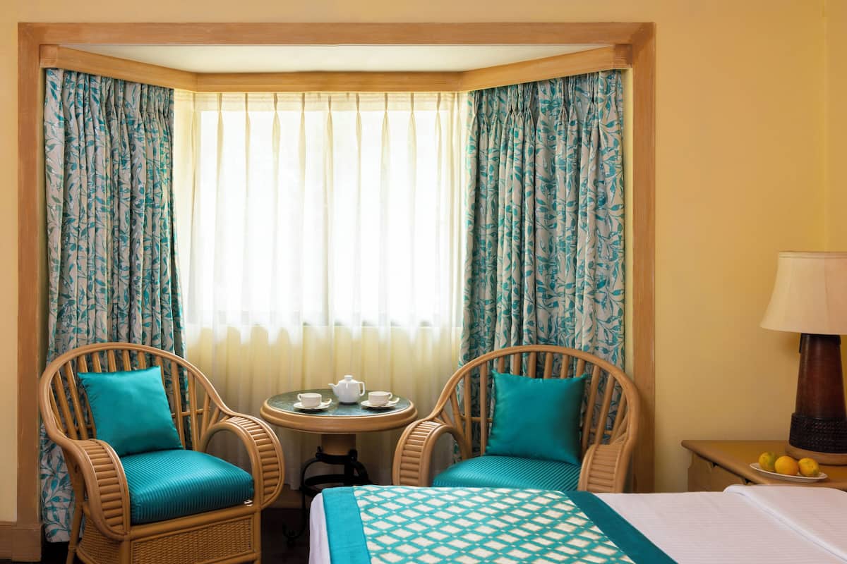 Ein Hotelzimmer mit einem Bett, zwei Korbstühlen, einem kleinen runden Tisch mit Teekanne und Tassen sowie einer Lampe auf einem Nachttisch.