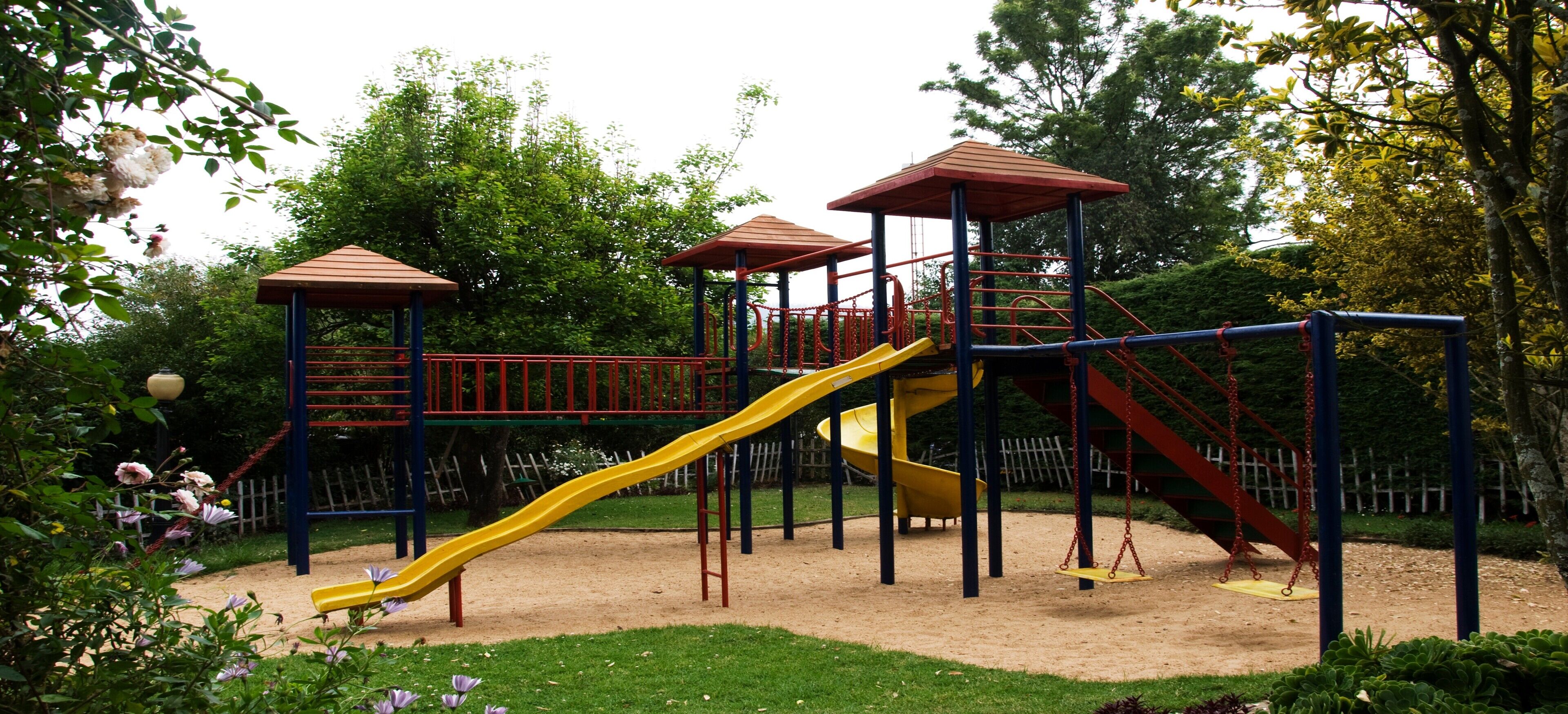 Un parque infantil con un tobogán amarillo, estructuras de escalada rojas y azules, y zonas cubiertas.