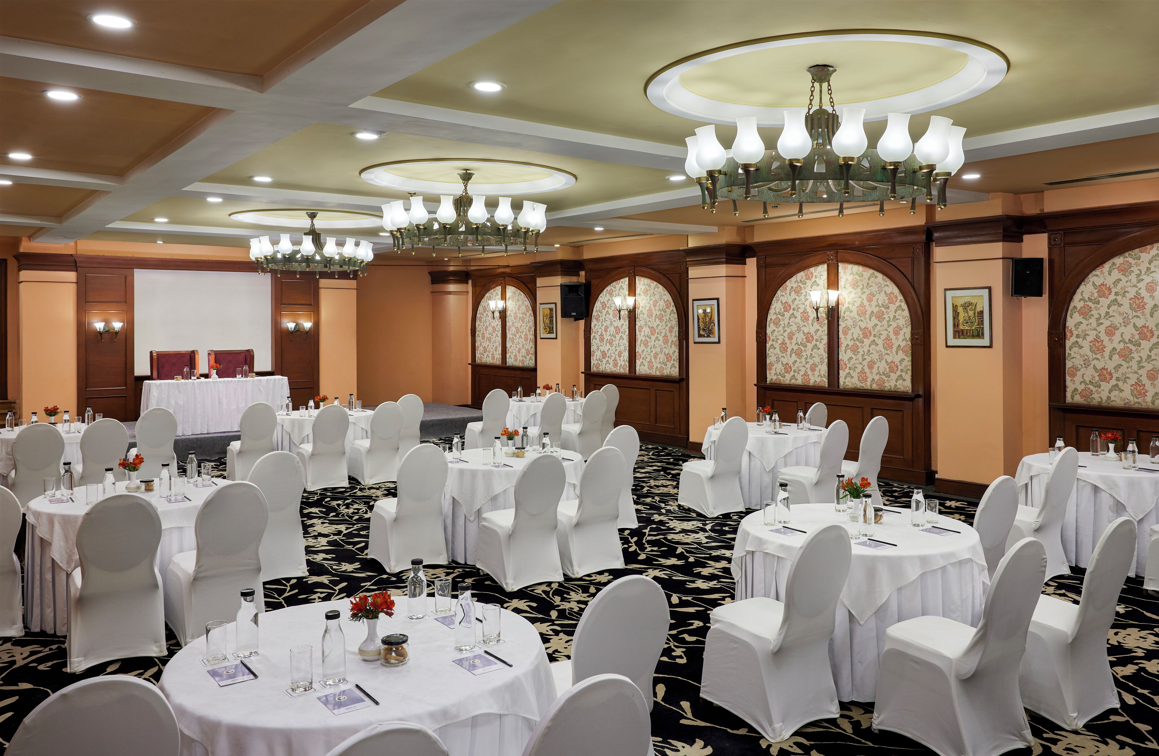 A banquet hall with round tables set for events, white chairs, chandeliers, and floral wallpaper.