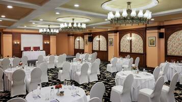 A banquet hall with round tables set for events, white chairs, chandeliers, and floral wallpaper.