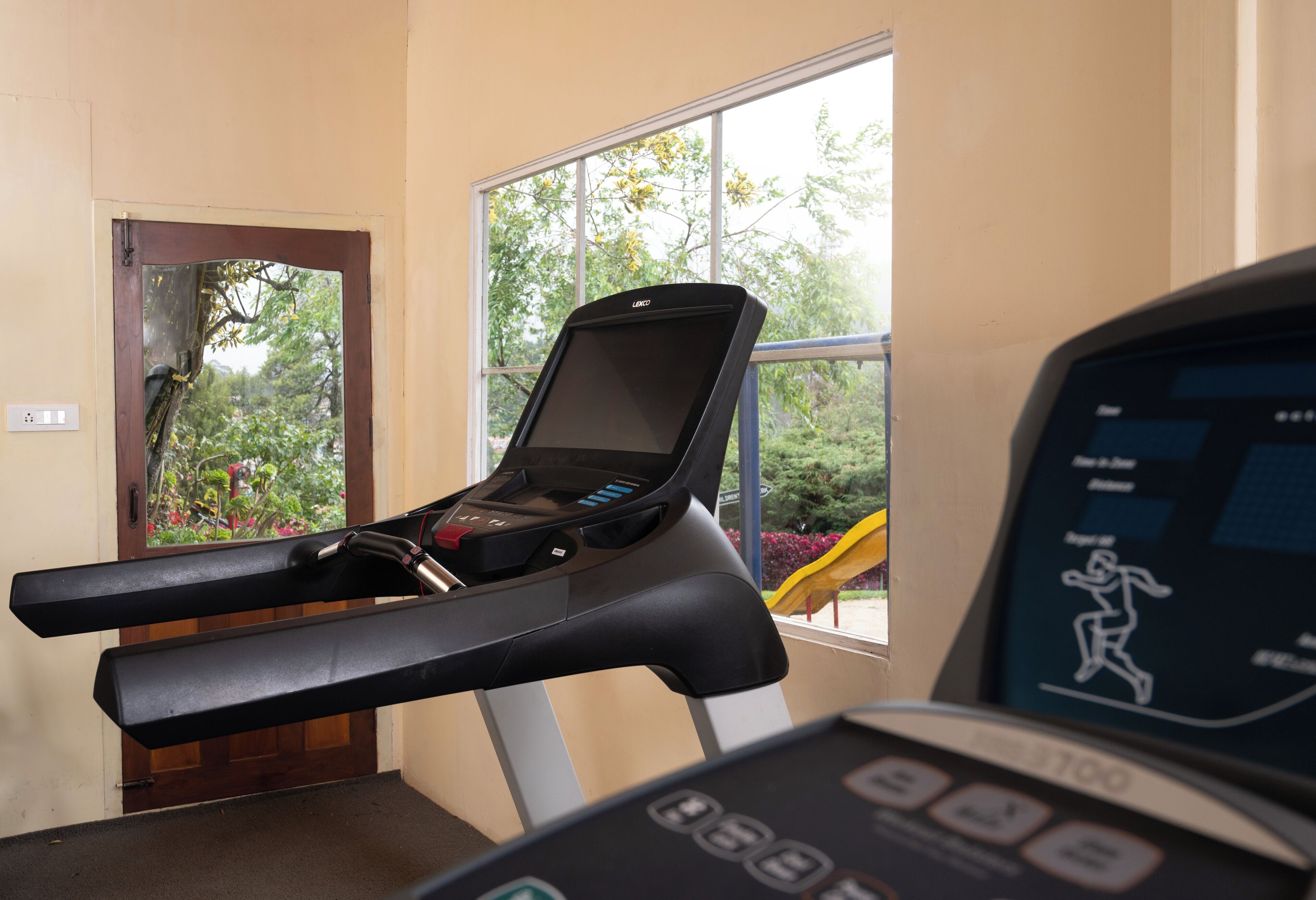 Esteira com visor digital, colocada perto de uma janela com vista para vegetação e um escorrega amarelo lá fora.