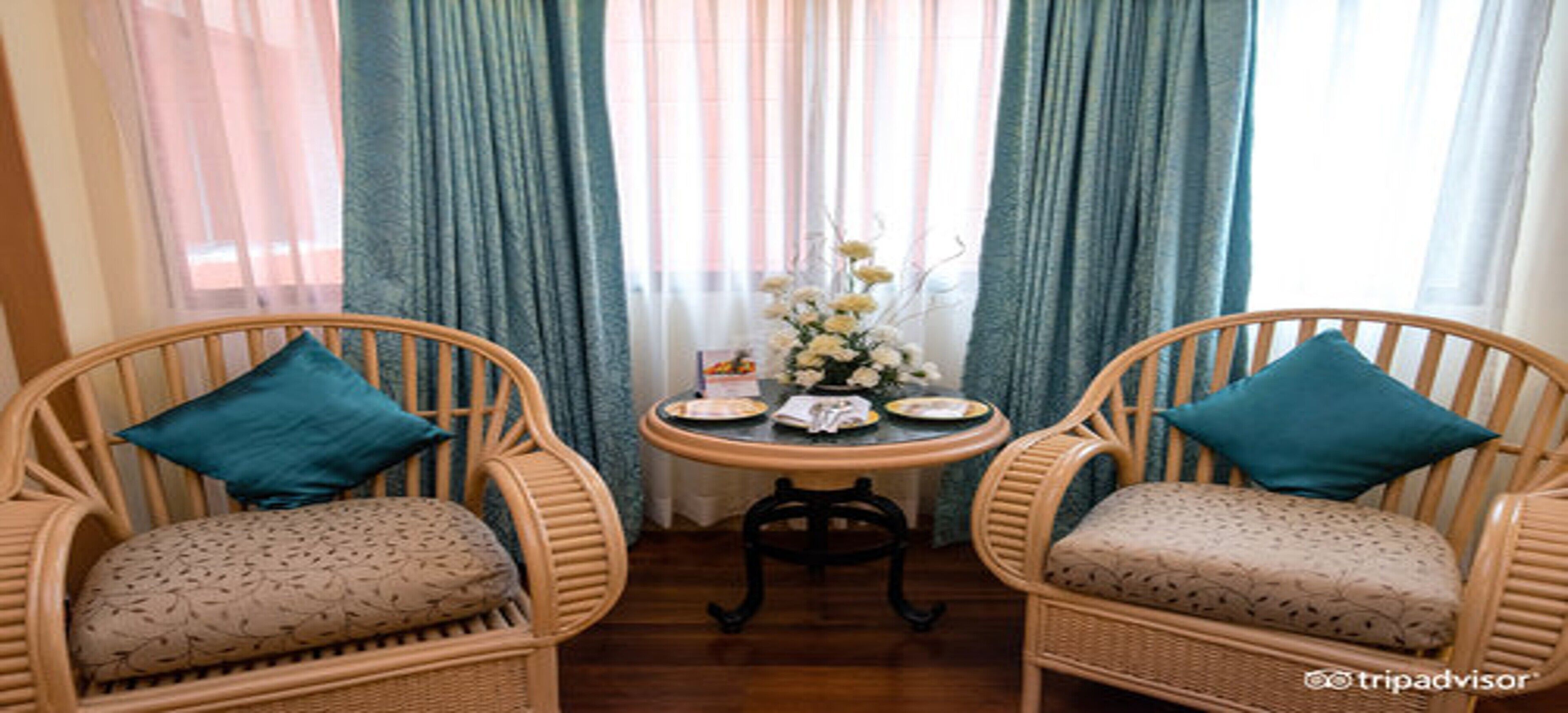 Une pièce avec deux fauteuils en osier dotés de coussins turquoise, une petite table ronde avec des assiettes et un vase de fleurs, et des rideaux bleus.