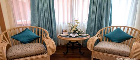 Une pièce avec deux fauteuils en osier dotés de coussins turquoise, une petite table ronde avec des assiettes et un vase de fleurs, et des rideaux bleus.
