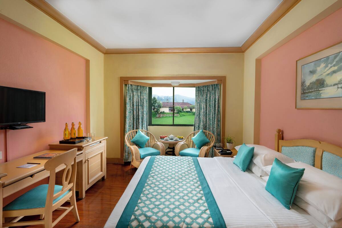 Ein Hotelzimmer mit einem Bett, einem Schreibtisch, zwei Stühlen, einem Fernseher und einem Fenster mit Blick auf ein Gebäude und Grünflächen.