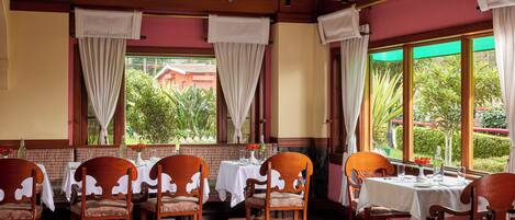 A dining area with wooden chairs and tables, large windows with curtains, and a view of greenery outside.