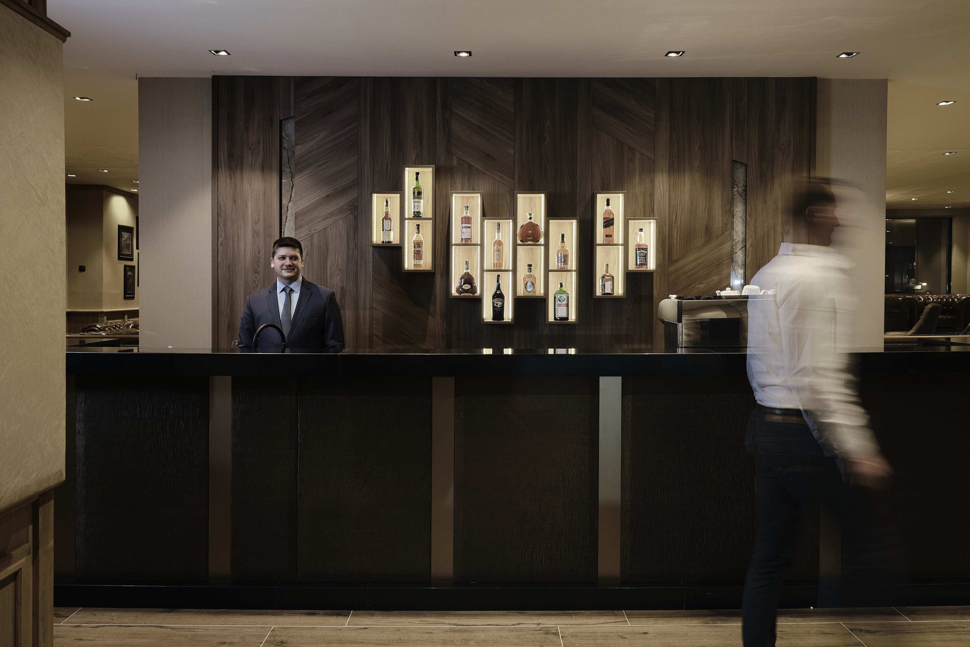 Zona de recepción de un hotel con un panel de madera, mostrador de recepción y una exhibición de botellas de licor.