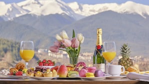 Une table de petit-déjeuner dressée avec des viennoiseries, des fruits et des boissons, le tout mis en valeur par un paysage montagneux en toile de fond.