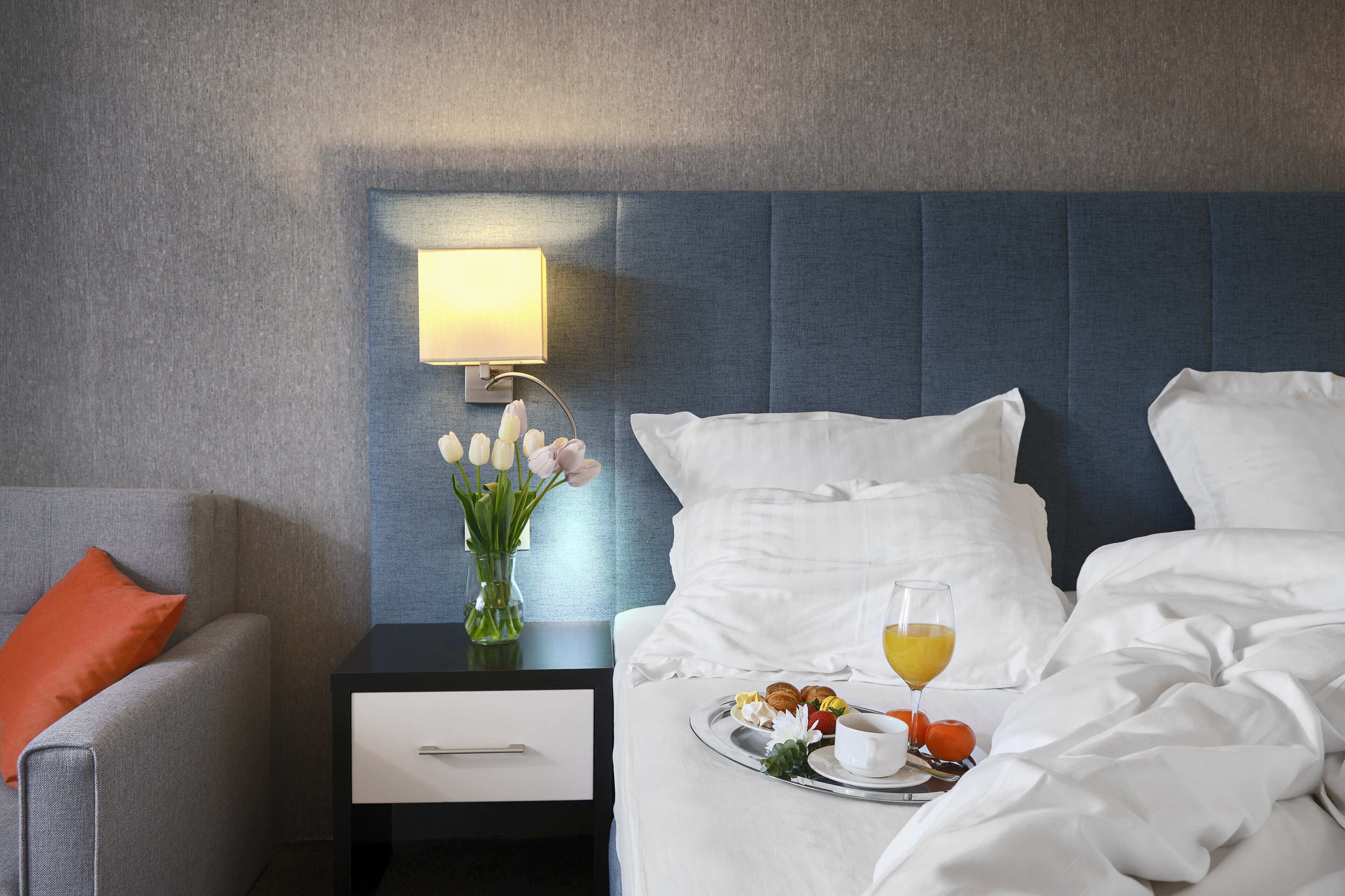 A hotel room with a bed, a nightstand with a lamp, a vase with flowers, and a tray with breakfast items.