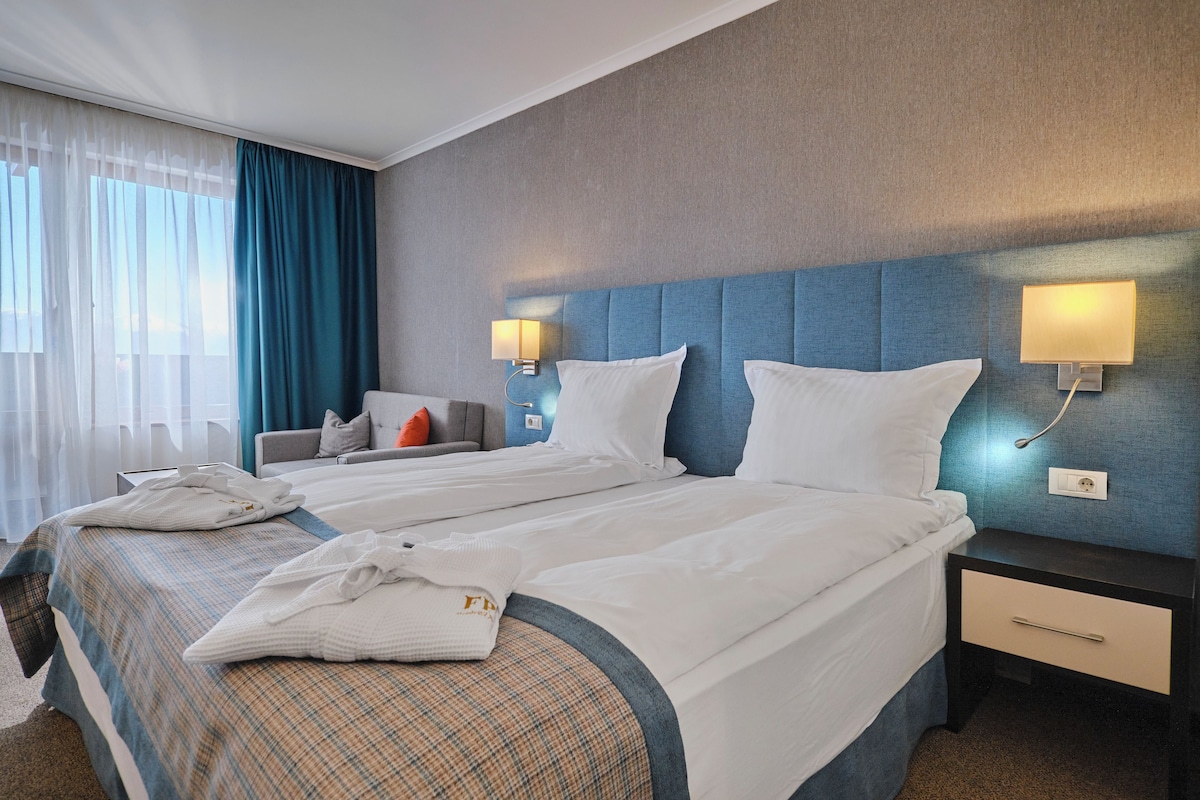 A hotel room with two beds, a nightstand with a lamp, a sofa, and a window with curtains.