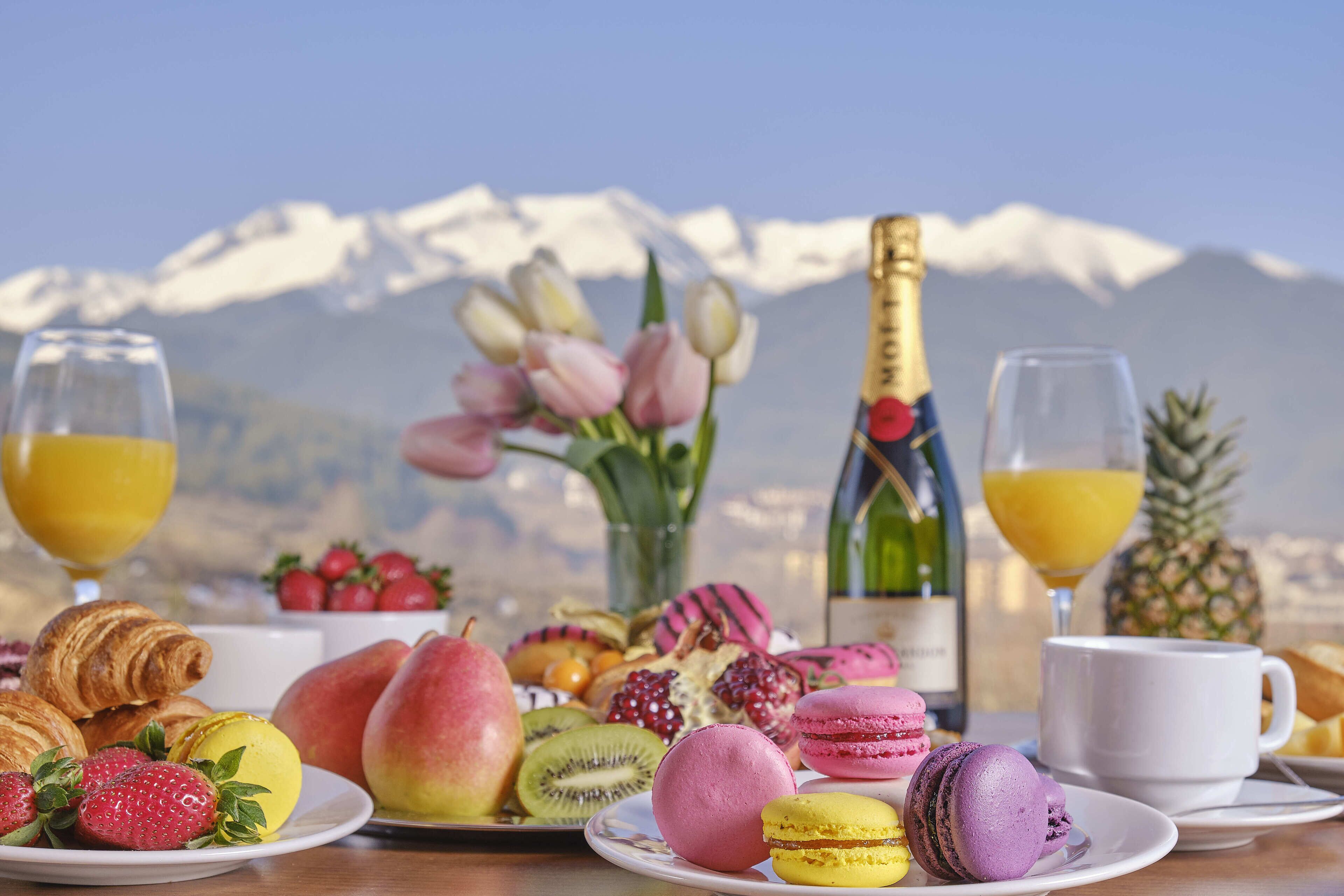 A breakfast table with pastries, fruits, and drinks, set against a mountain backdrop.