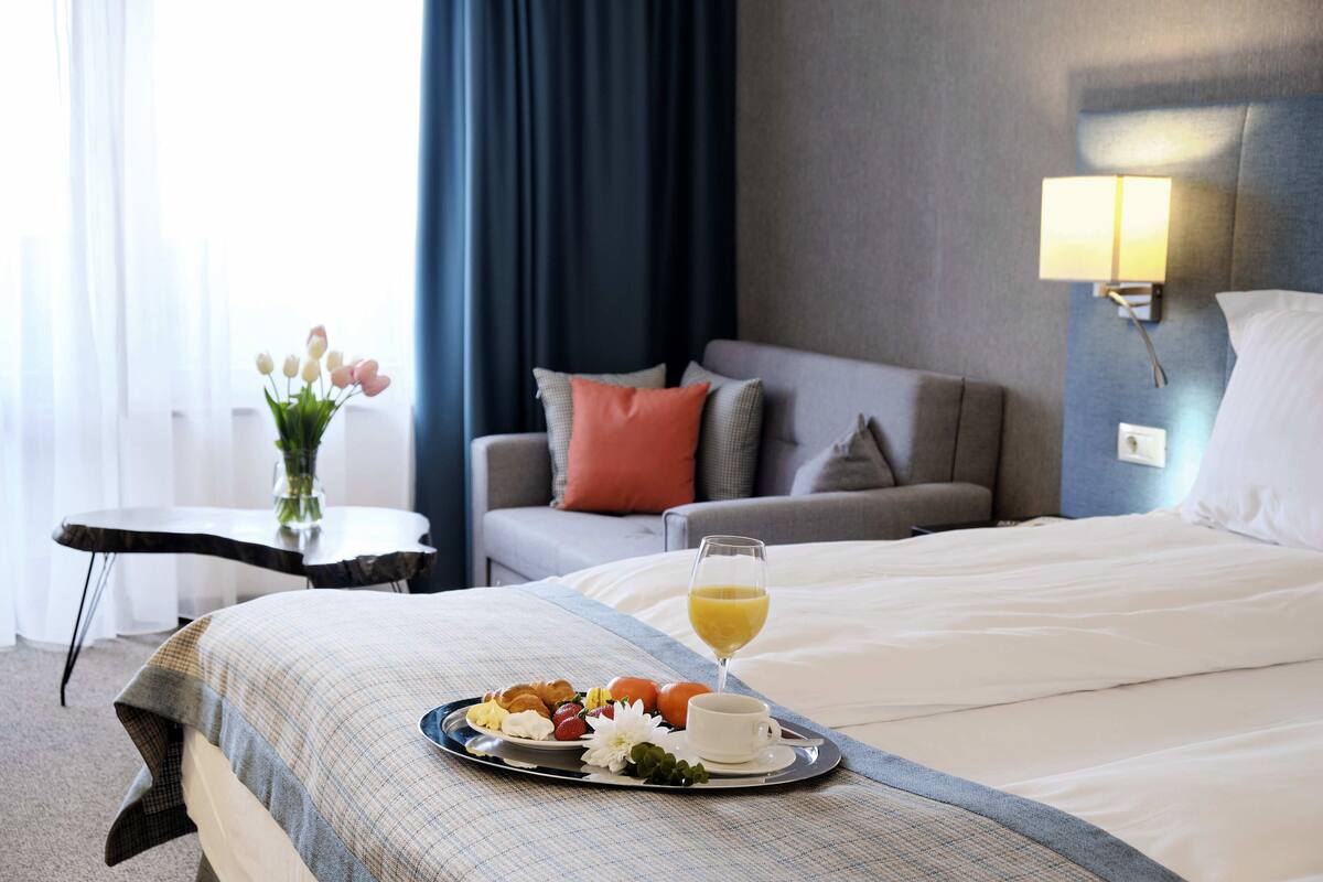 A hotel room with a bed, a bedside table with a vase of flowers, a tray with breakfast items, and a lamp on the wall.
