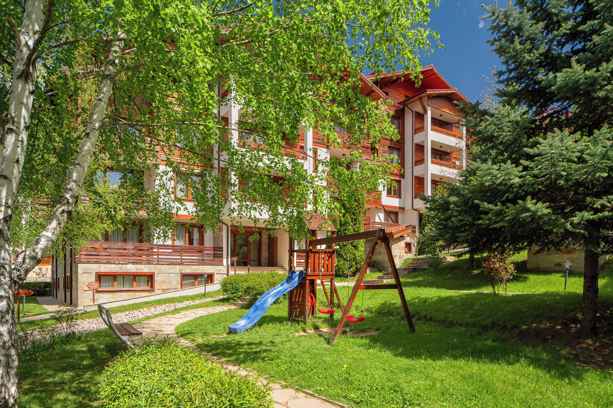 Un parque infantil con tobogán, columpios y una estructura para escalar frente a un edificio con balcones y un tejado rojo.