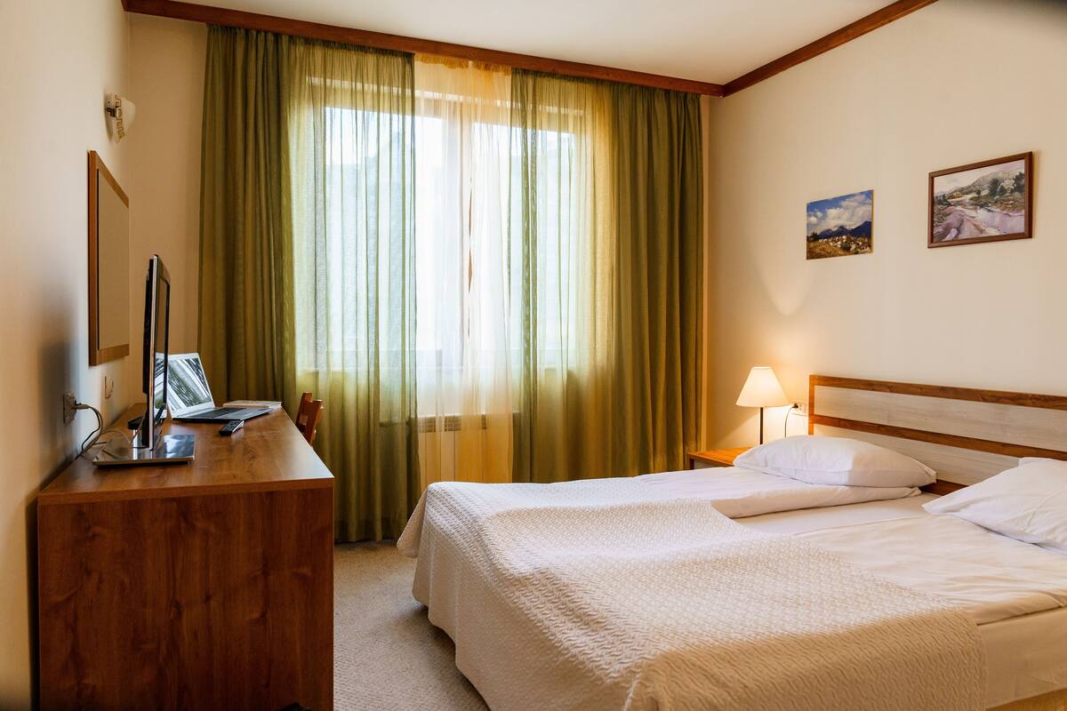 A hotel room with a bed, a desk with a computer, a lamp, and two paintings on the wall.