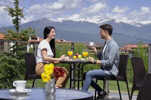 Dos personas sentadas en una mesa disfrutando del desayuno con frutas y jugo de naranja, con vistas a la montaña.