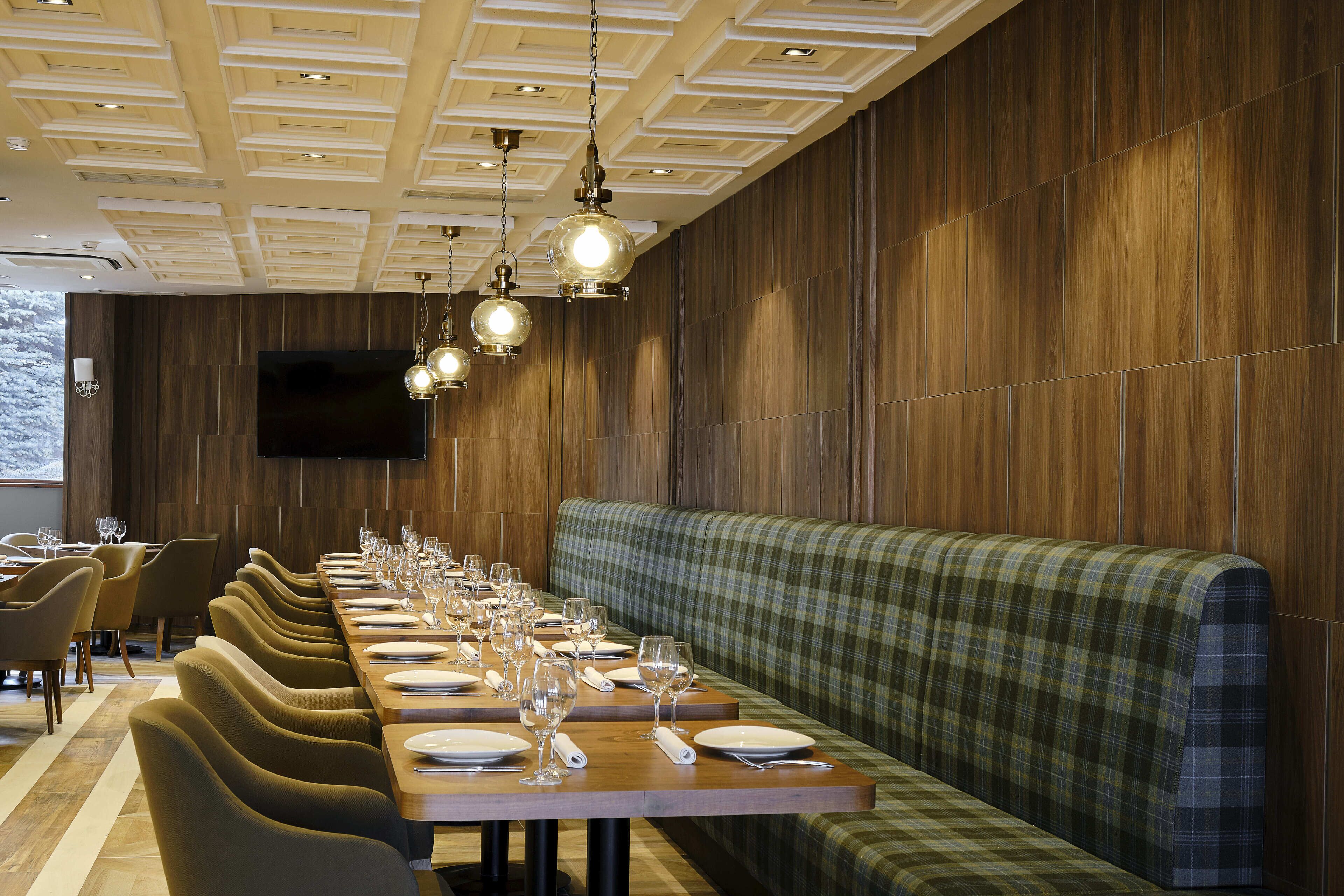 A restaurant with a long wooden table set for dining, a green plaid bench, and pendant lighting.
