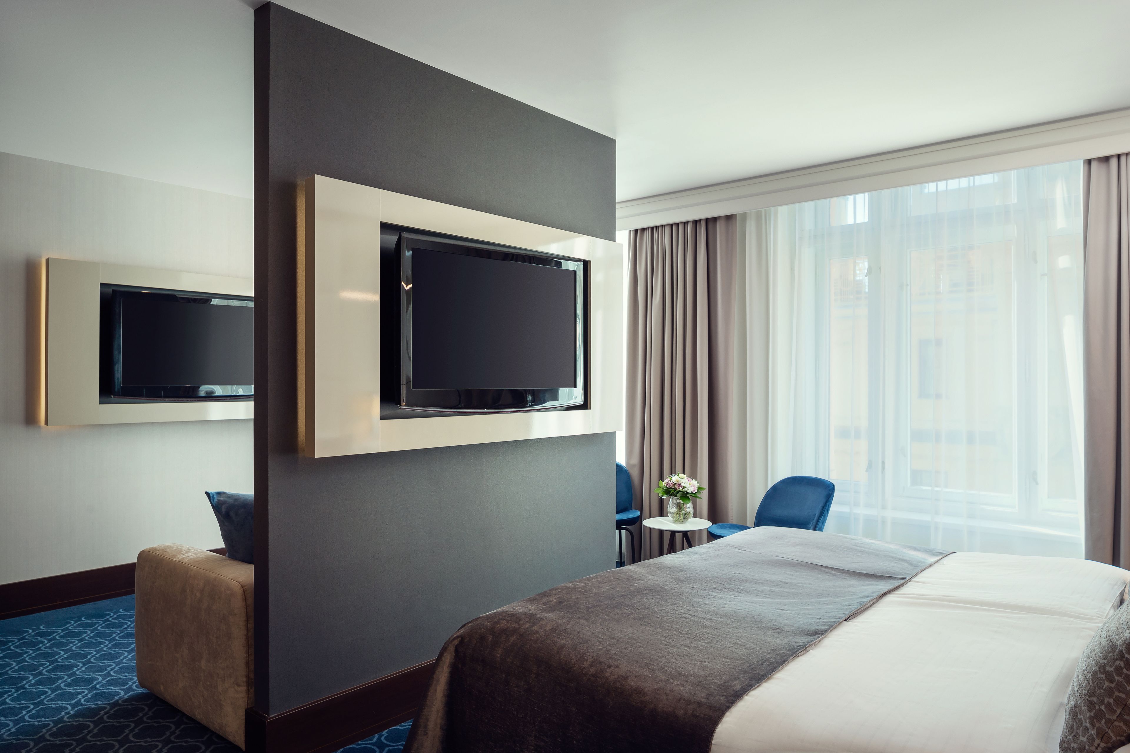 Een hotelkamer met een groot bed, een flatscreen televisie aan de muur, een kleintje tafeltje met een vaas, en een raam met gordijnen.