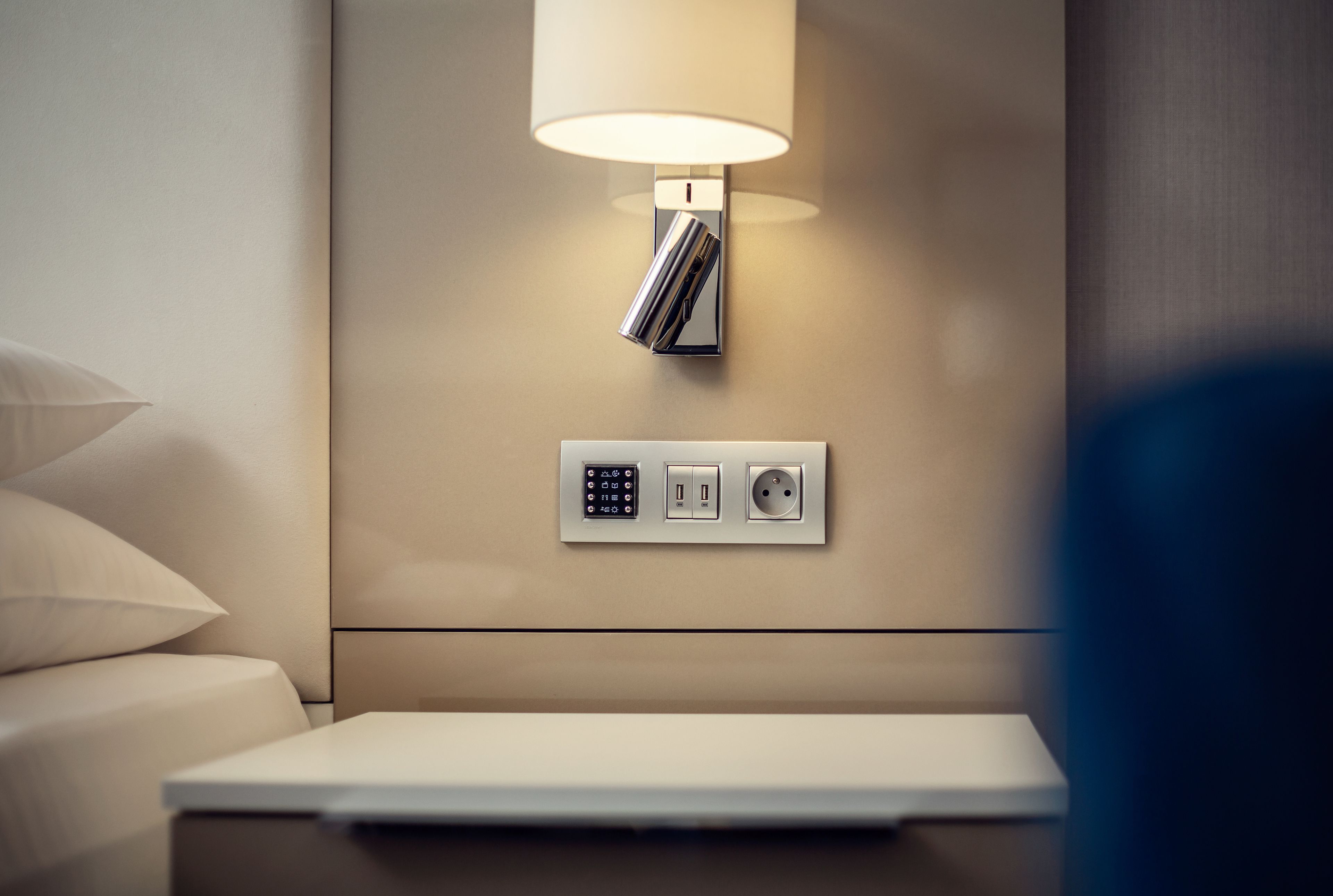 Een moderne hotelkamer met nachtkastje, lamp en een stopcontact op de muur.