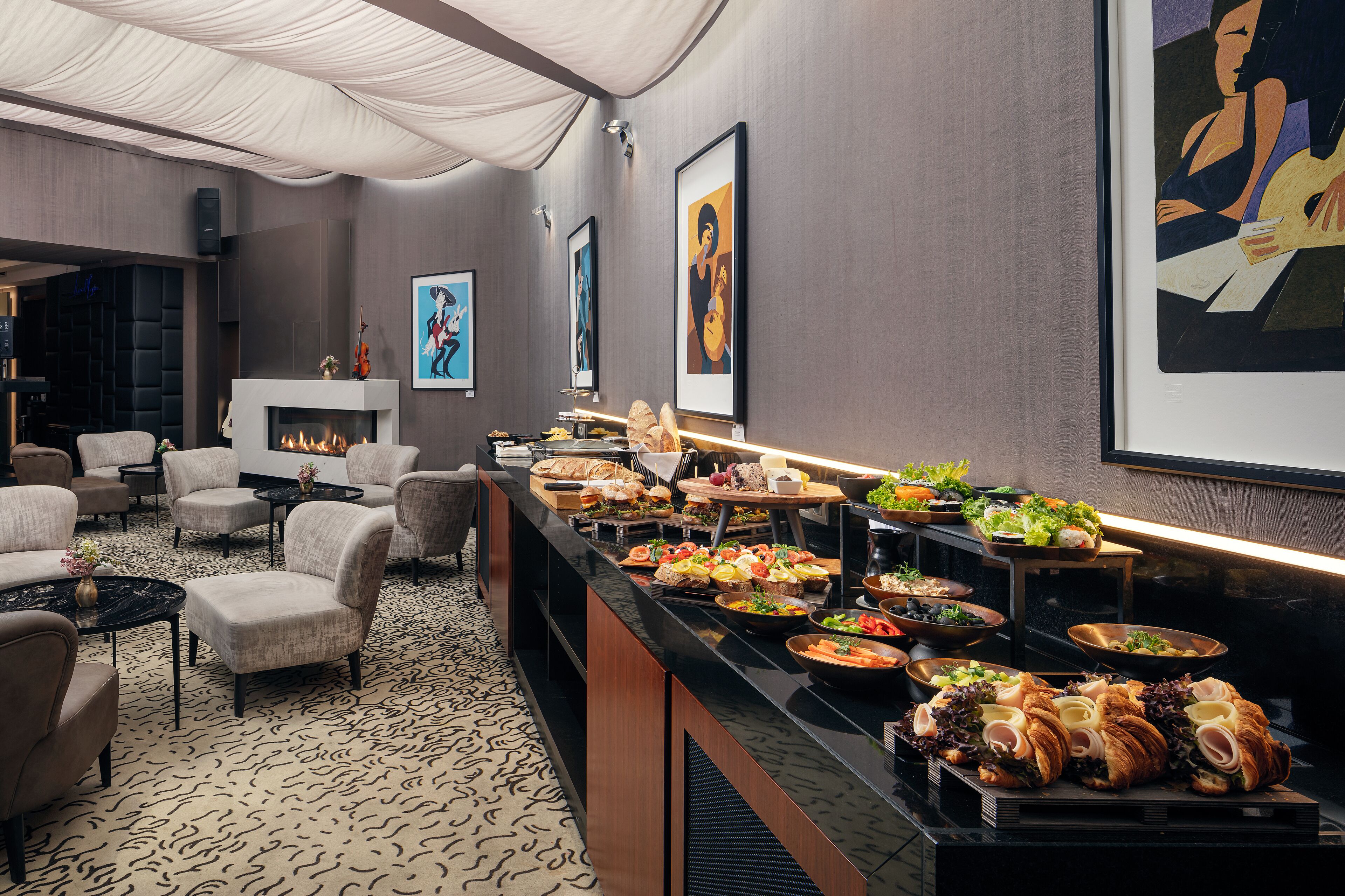 Disposição de buffet com diversos alimentos, lareira e área de estar com cadeiras e mesas.