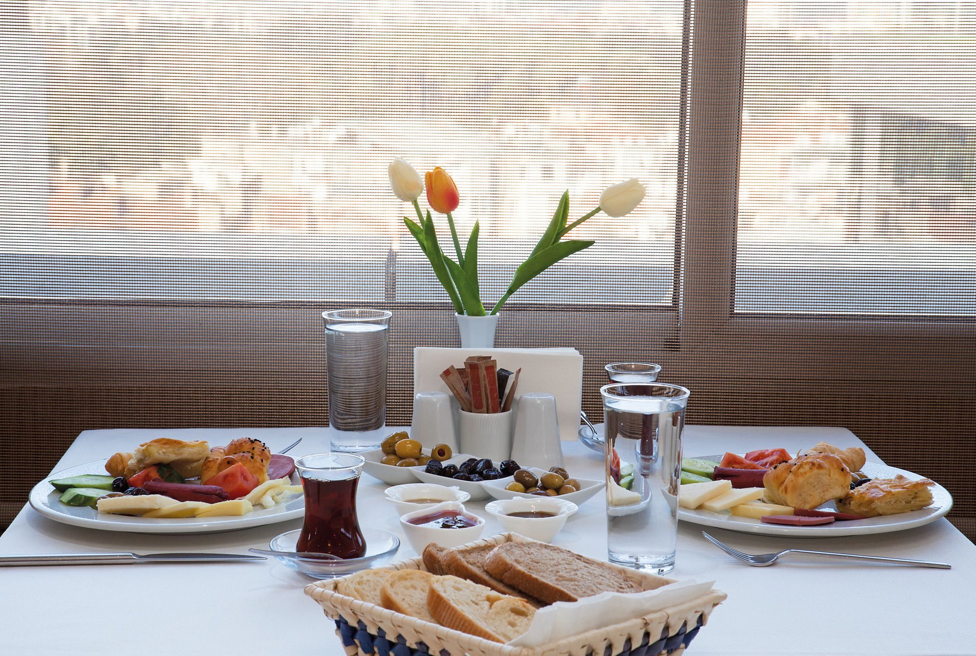 Ein Esstisch mit verschiedenen Speisen, darunter Brot, Früchte und Gebäck, sowie eine Vase mit Tulpen.