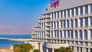 A multi-story hotel with a prominent 'Enjoy' sign, featuring balconies with railings, a view of the sea, and a desert landscape in the background. - Brown Dead Sea by Brown Hotels (Tamar)