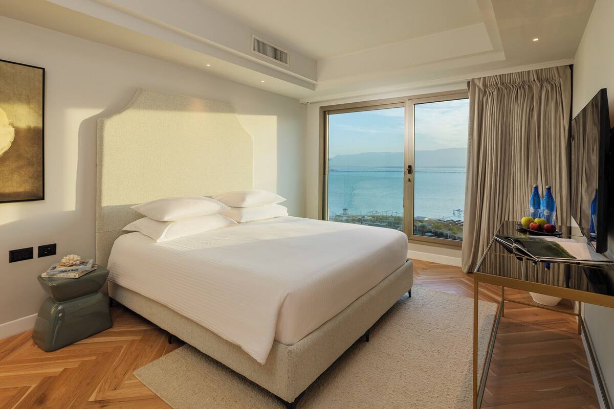 A modern hotel room with a large bed, a view of the sea, and a small table with bottles and fruit.