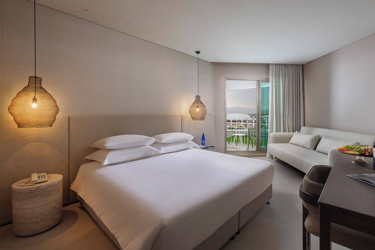 A hotel room with a bed, a sofa, a dining table with a bottle and glasses, a phone on a side table, and a balcony with a view.