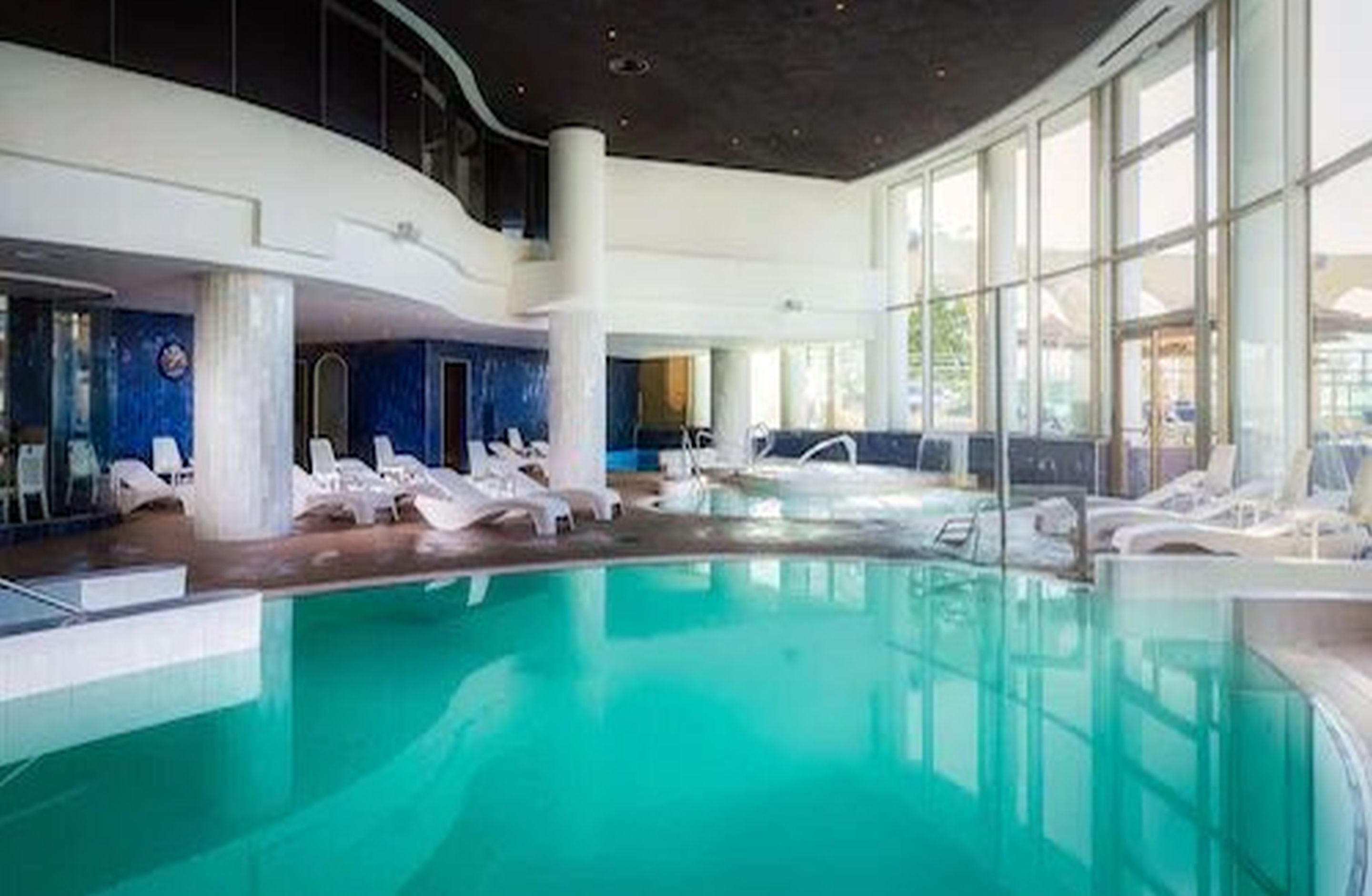 An indoor swimming pool with clear blue water, white lounge chairs, and large windows allowing natural light.